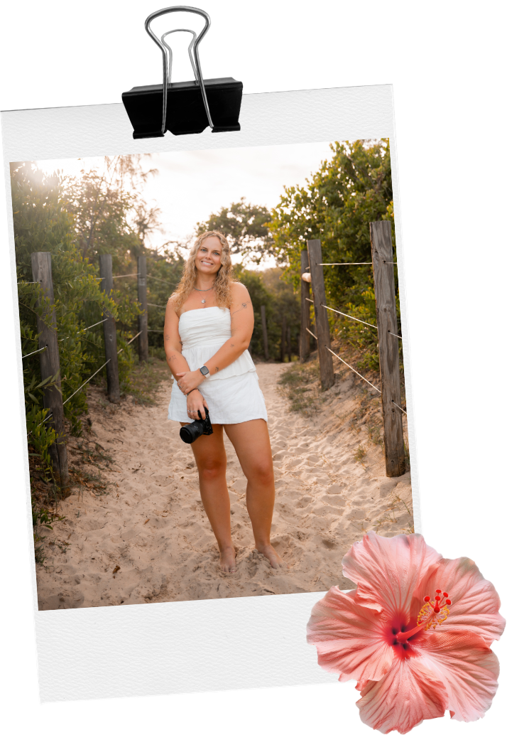 Young woman in white dress holding camera on sandy beach path, surrounded by wooden fences and greenery, with pink hibiscus flower.