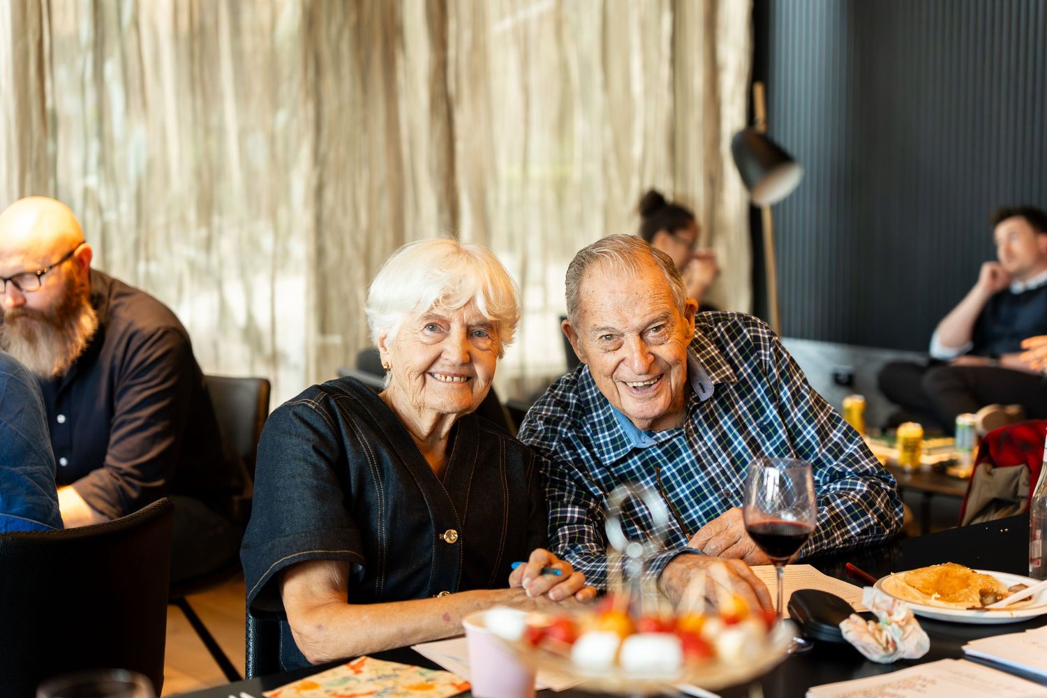 Elderly couple smiling together at a restaurant table with wine and food, enjoying a warm dining experience.