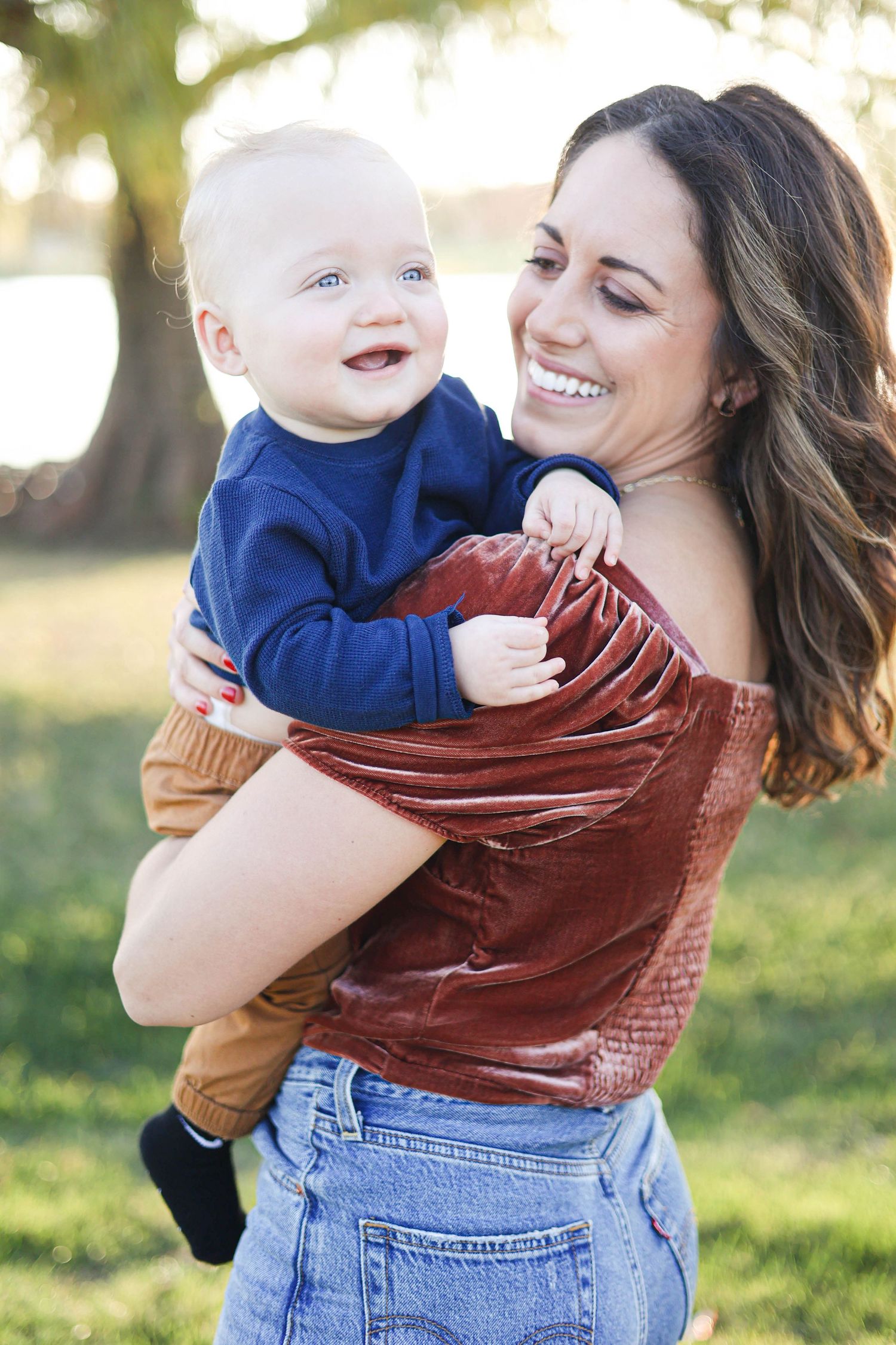 Mother holding infant son, smiling and laughing in the park during golden hour.