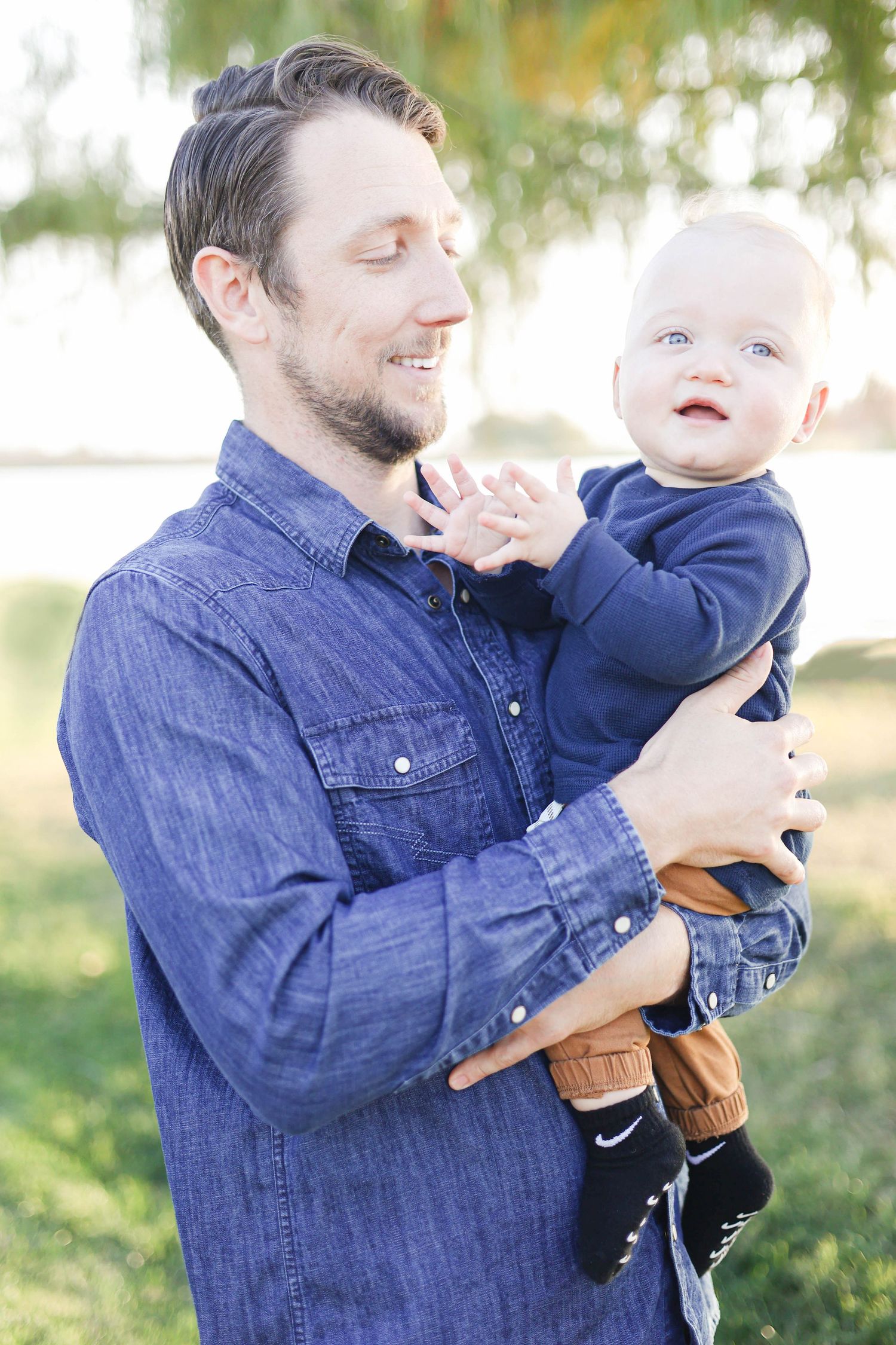 Father holding infant son, smiling and laughing in the park during golden hour.
