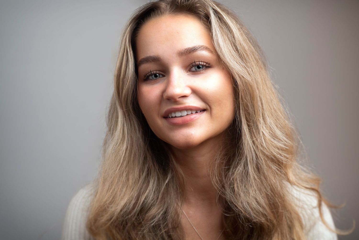 Warm portrait of a young woman with long blonde hair smiling against neutral background.