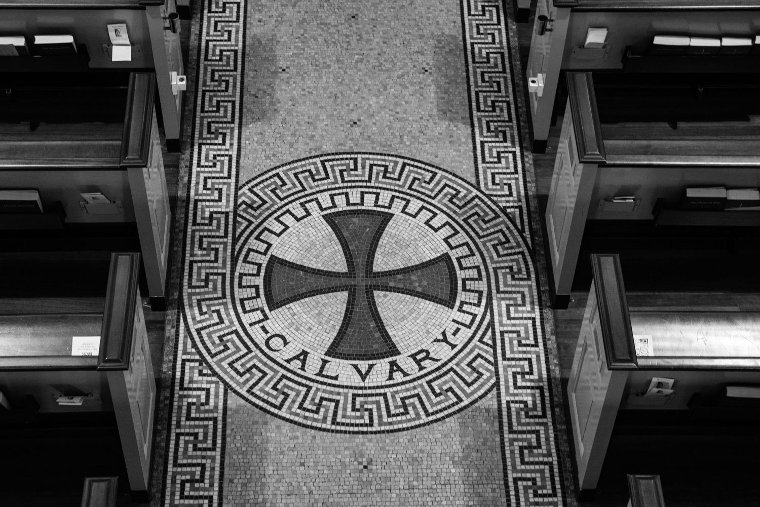Decorative floor mosaic featuring a Maltese cross surrounded by a Greek key pattern between wooden pews.John's Episcopal Church Georgetown