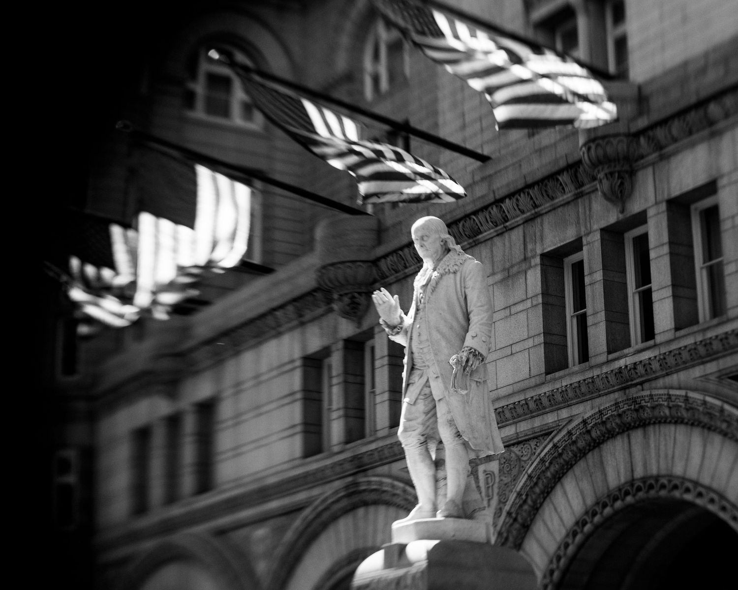 Black and white photo of a historic statue before a grand building with American flags waving in the background.