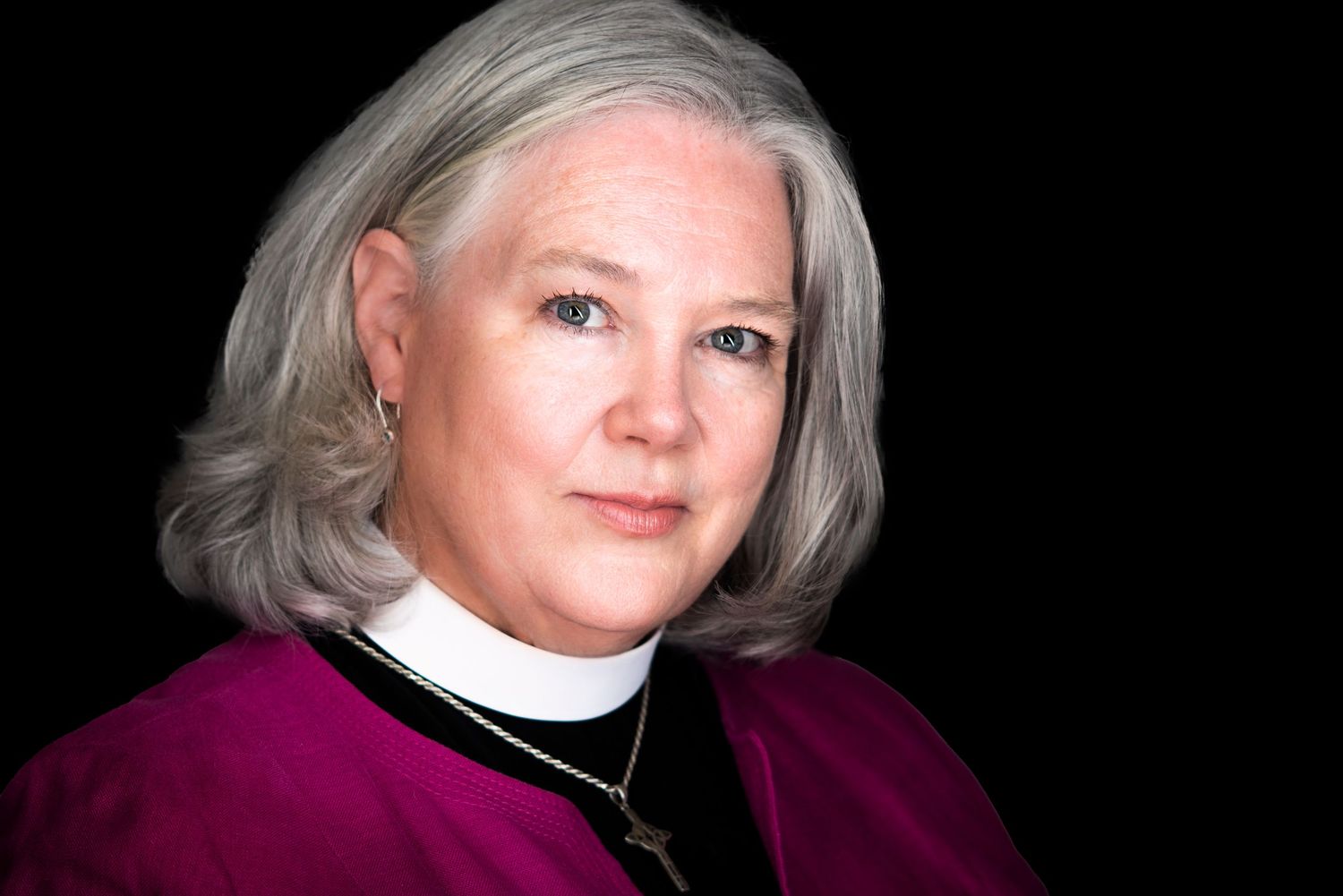 Portrait of a female clergy member wearing a magenta robe and clerical collar against black background.