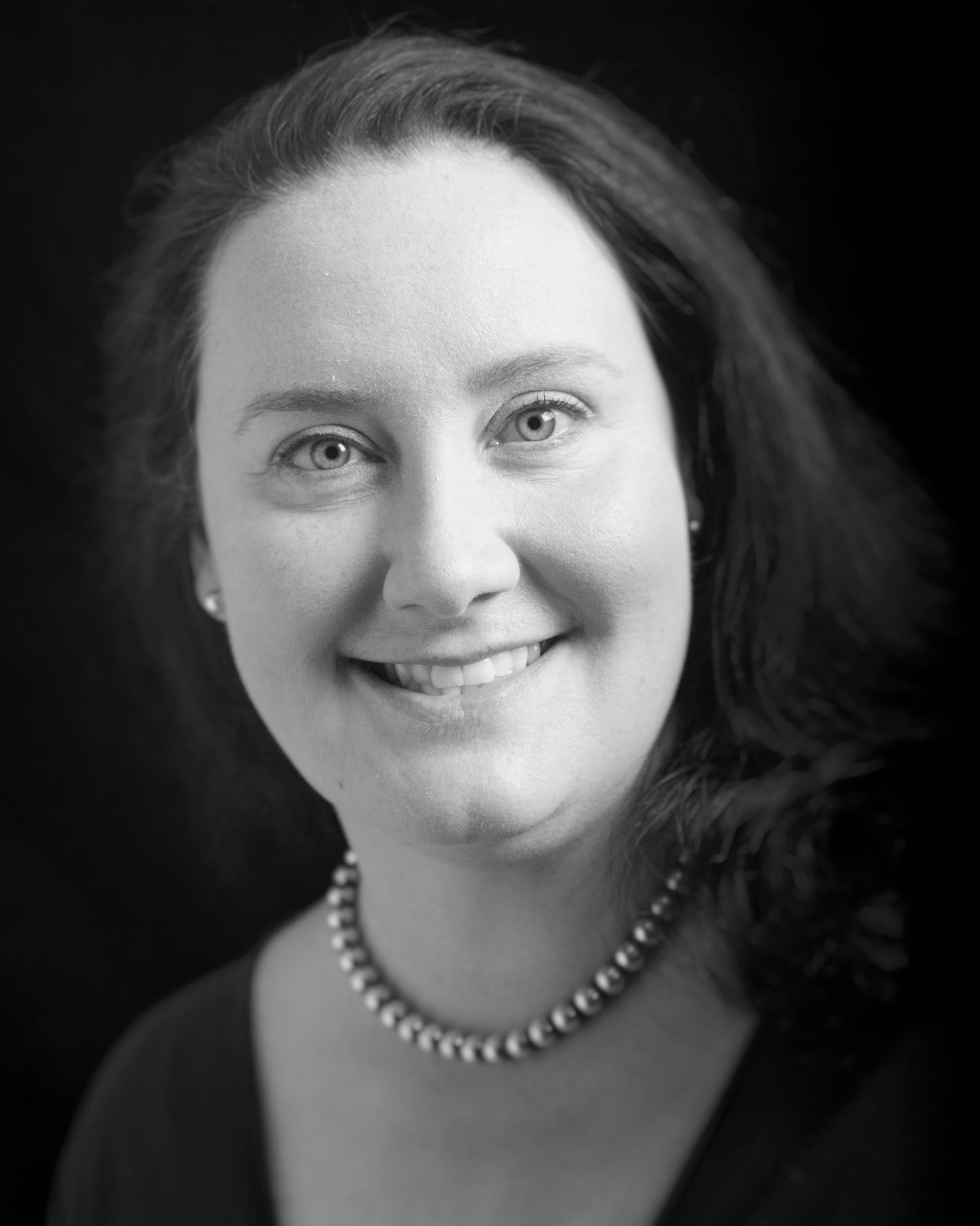 Professional headshot of a smiling woman wearing a black top and pearl necklace against dark backdrop.