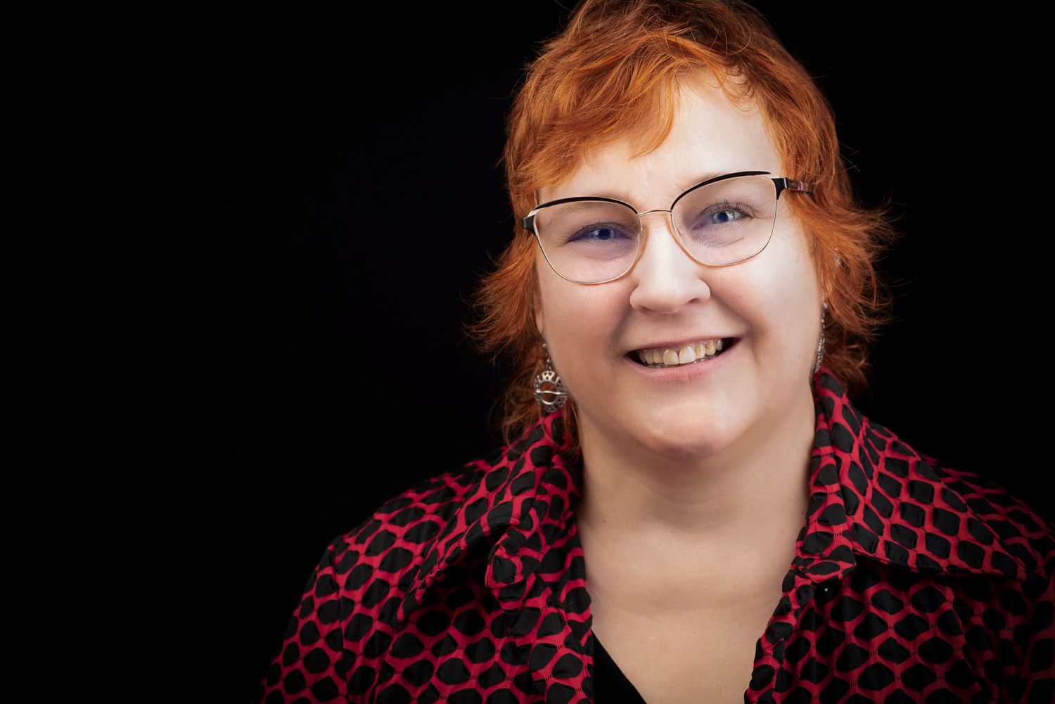 Portrait of a woman with red hair wearing glasses and a print top against dark background.