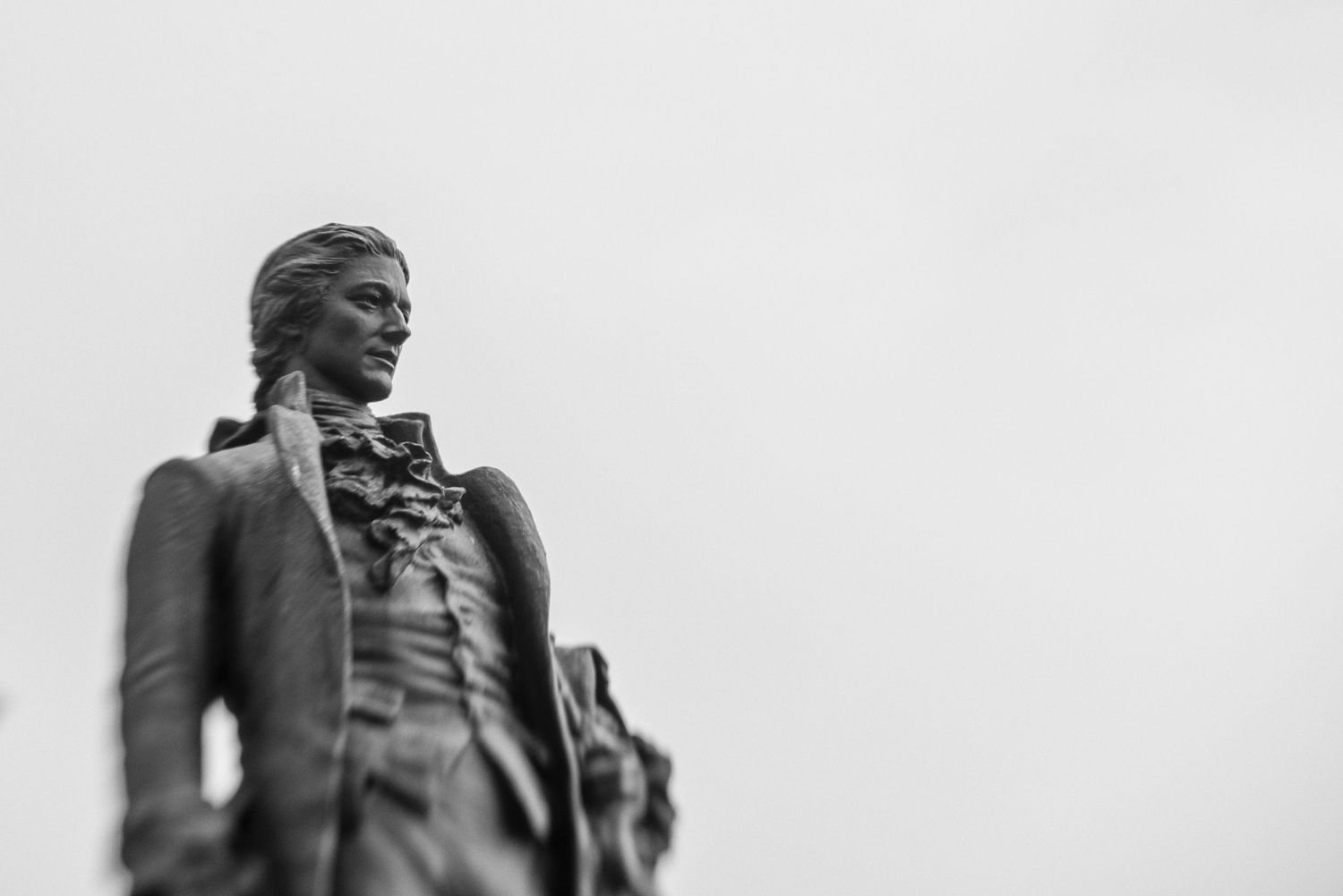 Black and white statue of a man in 18th-century attire standing against a cloudy sky.