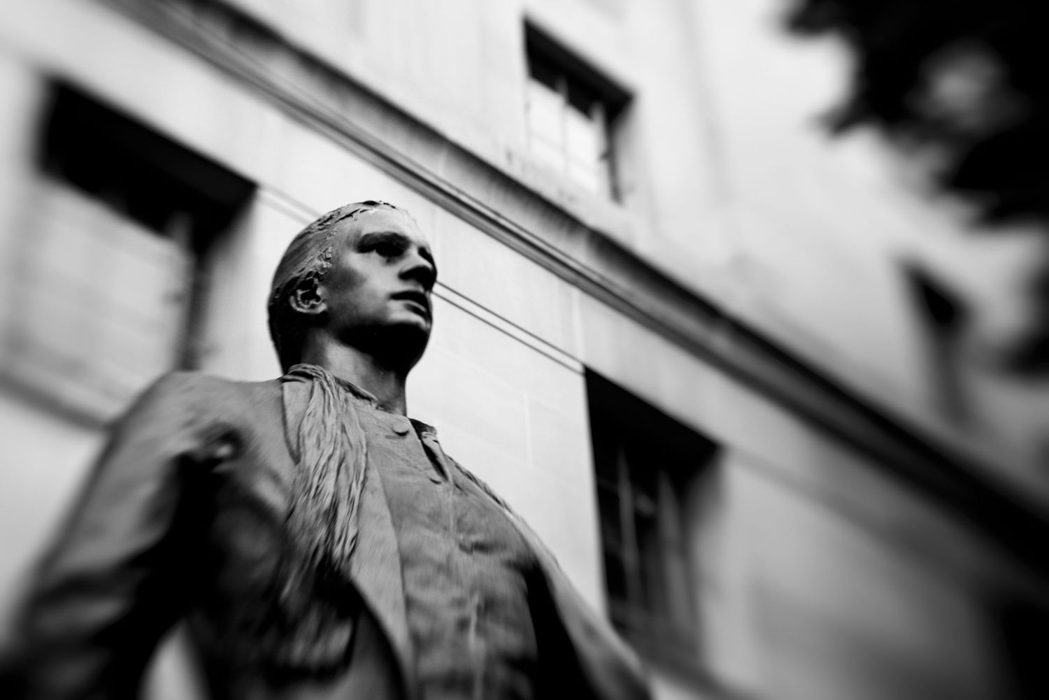 Black and white photo of a bronze statue of a man gazing upward, set against a building facade.