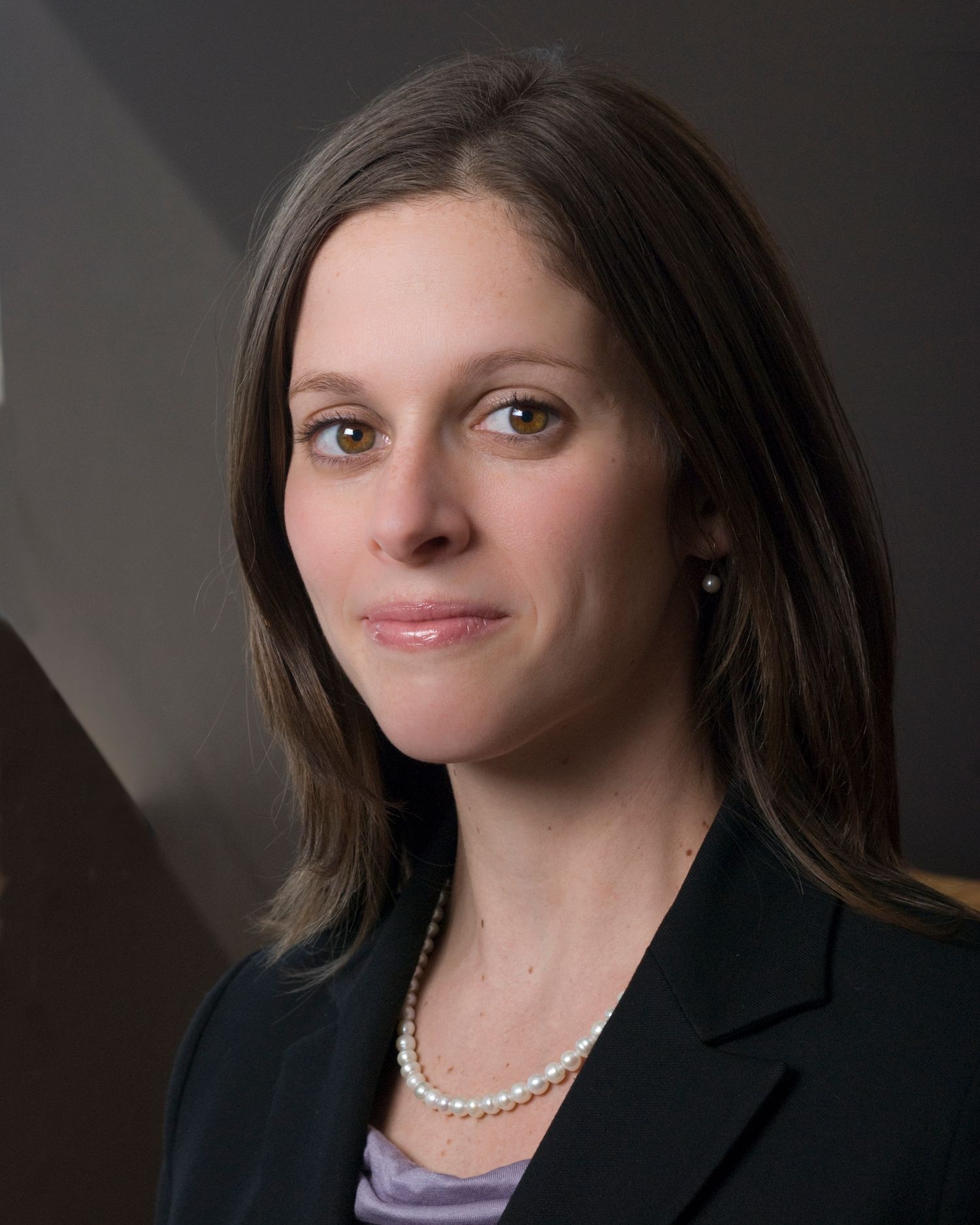 Professional headshot of a pwoman in business attire with pearl necklace against dark backdrop.