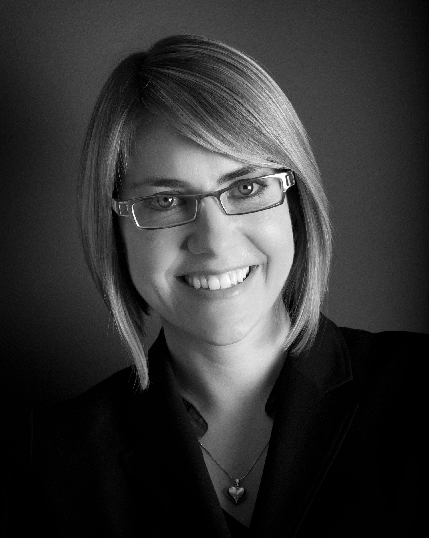 Black and white portrait of a woman featuring glasses and smile against dark backdrop.