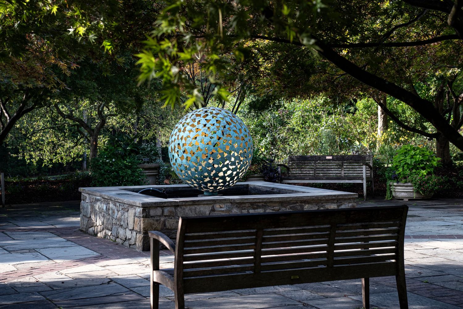A decorative blue metal sphere sculpture sits on a stone wall near a wooden bench in a peaceful garden setting.