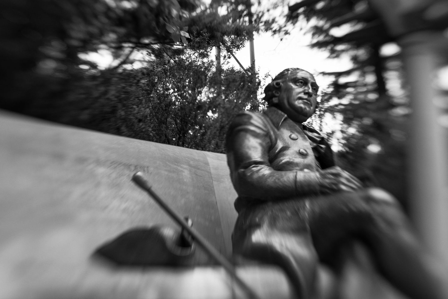 Black and white photo of a bronze statue seated on a bench, captured with a wide-angle lens outdoors.
