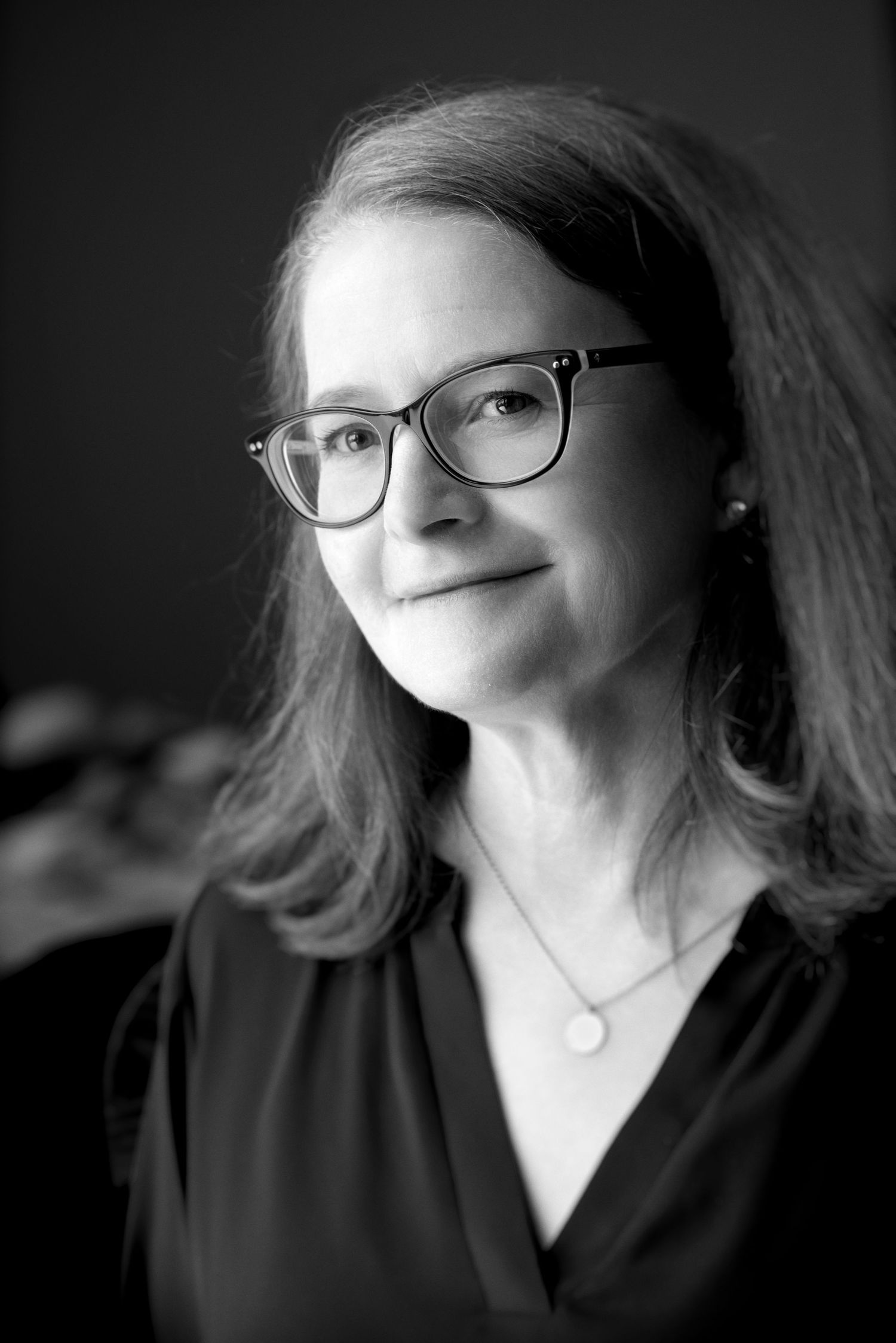 Professional headshots of womanin glasses with shoulder-length hair against dark backdrop.