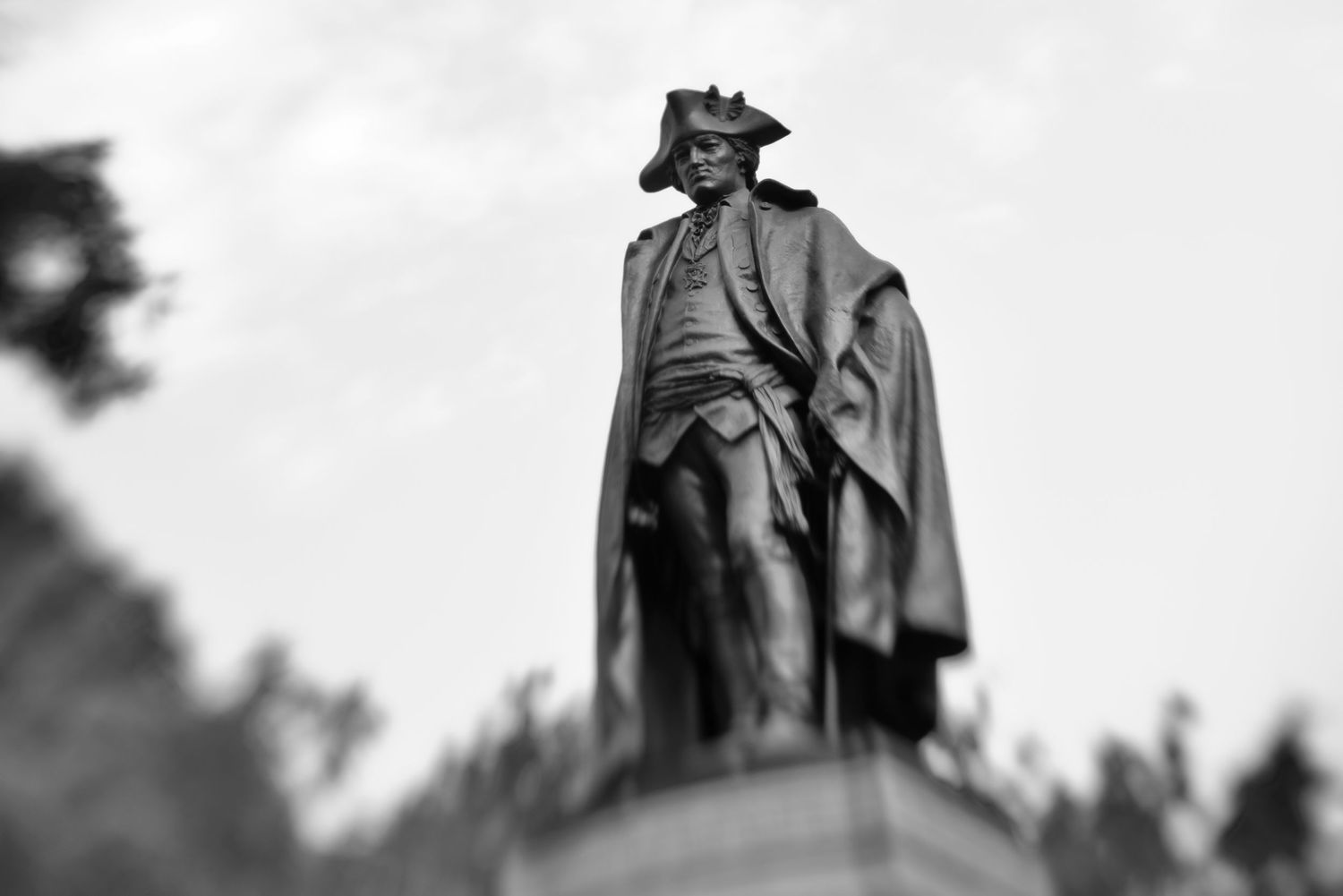 Black and white photo of a historic bronze statue of a military figure wearing a cape and tricorn hat against a cloudy sky.