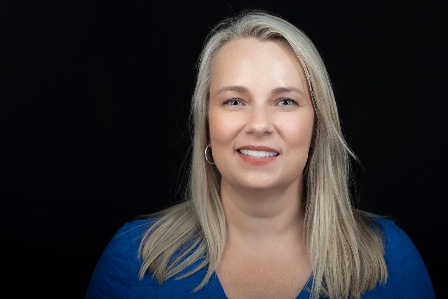 Professional headshot of a woman in blue top smiling against black background.