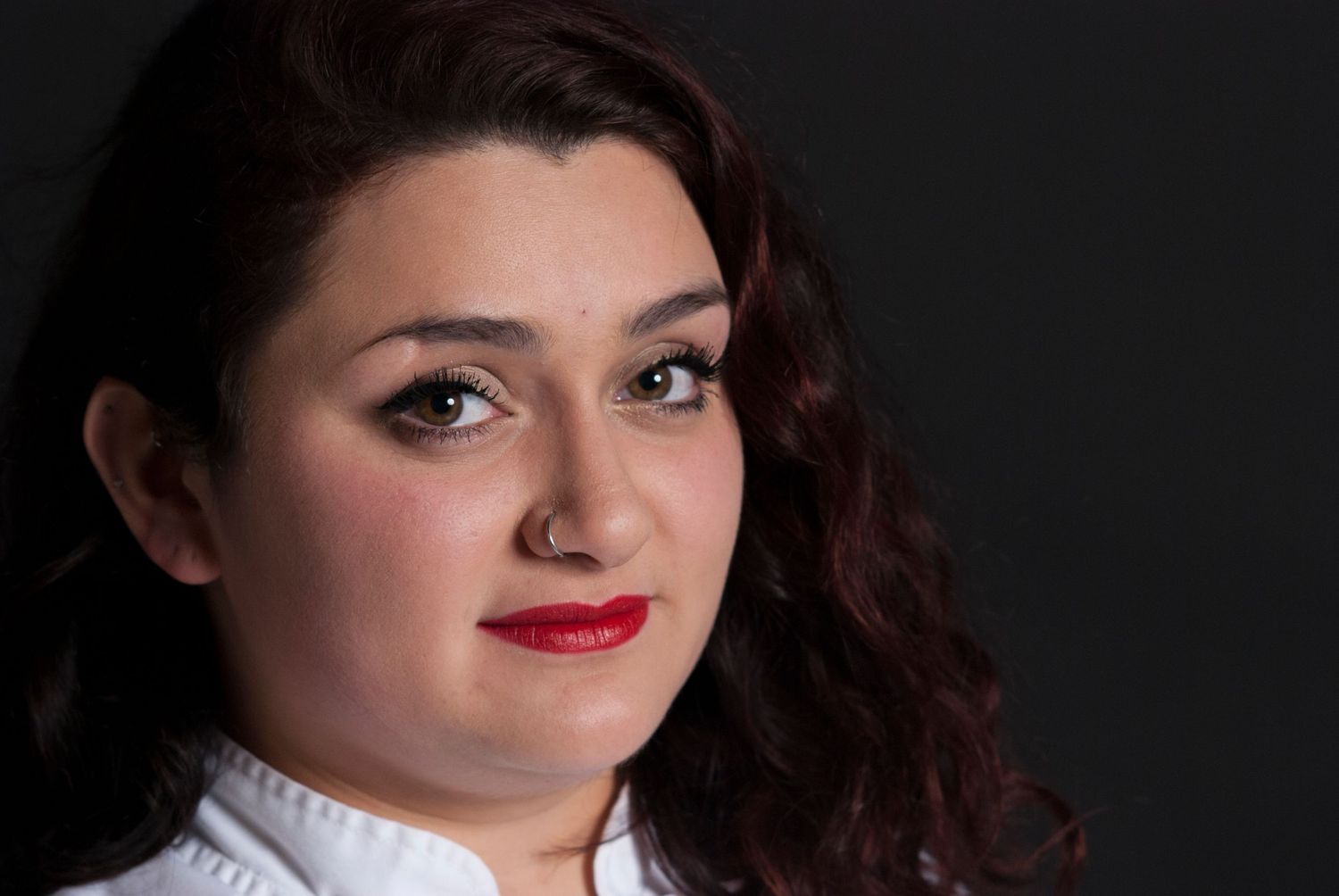 Headshot of young woman with dark curly hair, and bright red lipstick wearing a chef's jacket