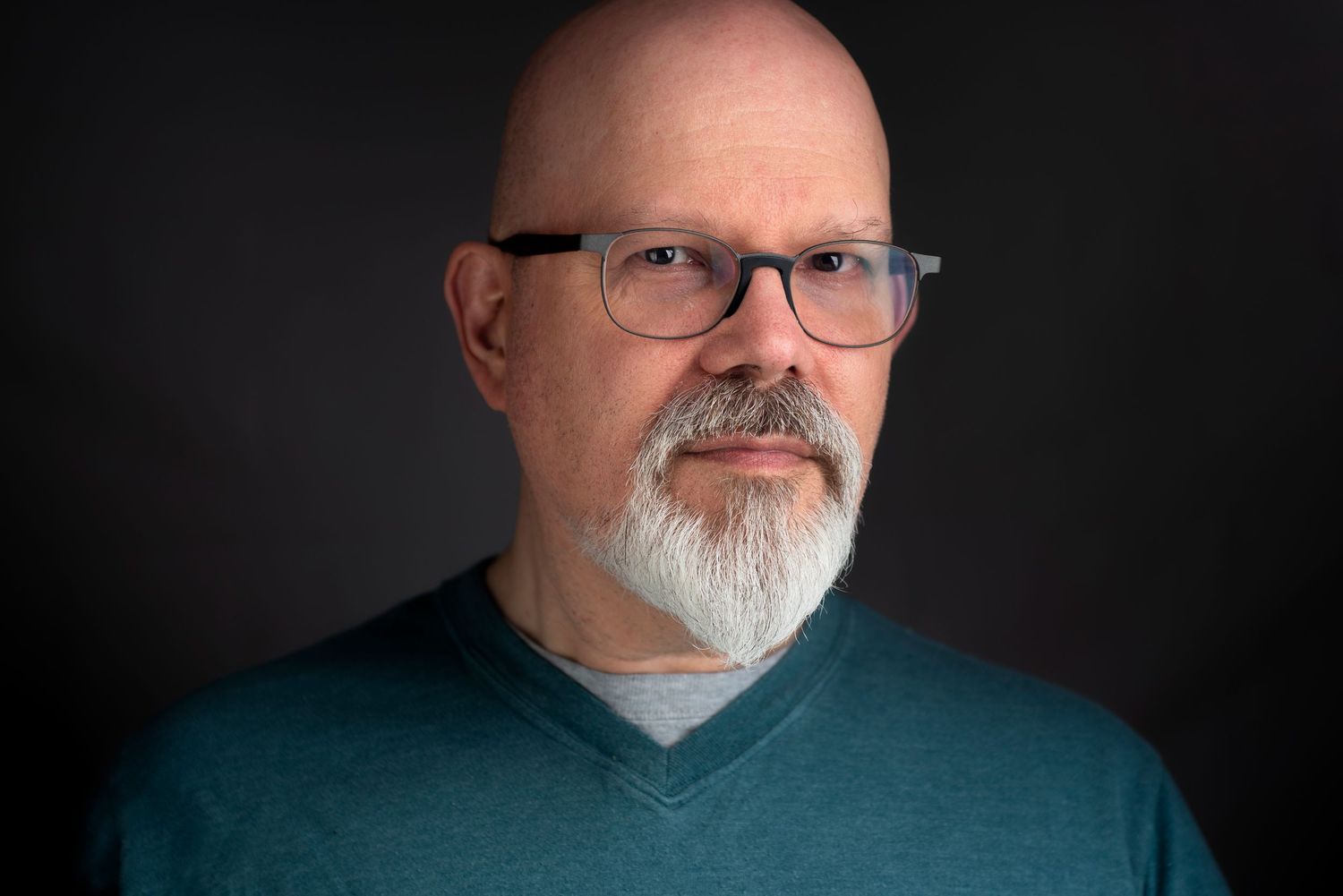 Professional portraits of a man with glasses, bald head, and grey beard against dark backdrop.