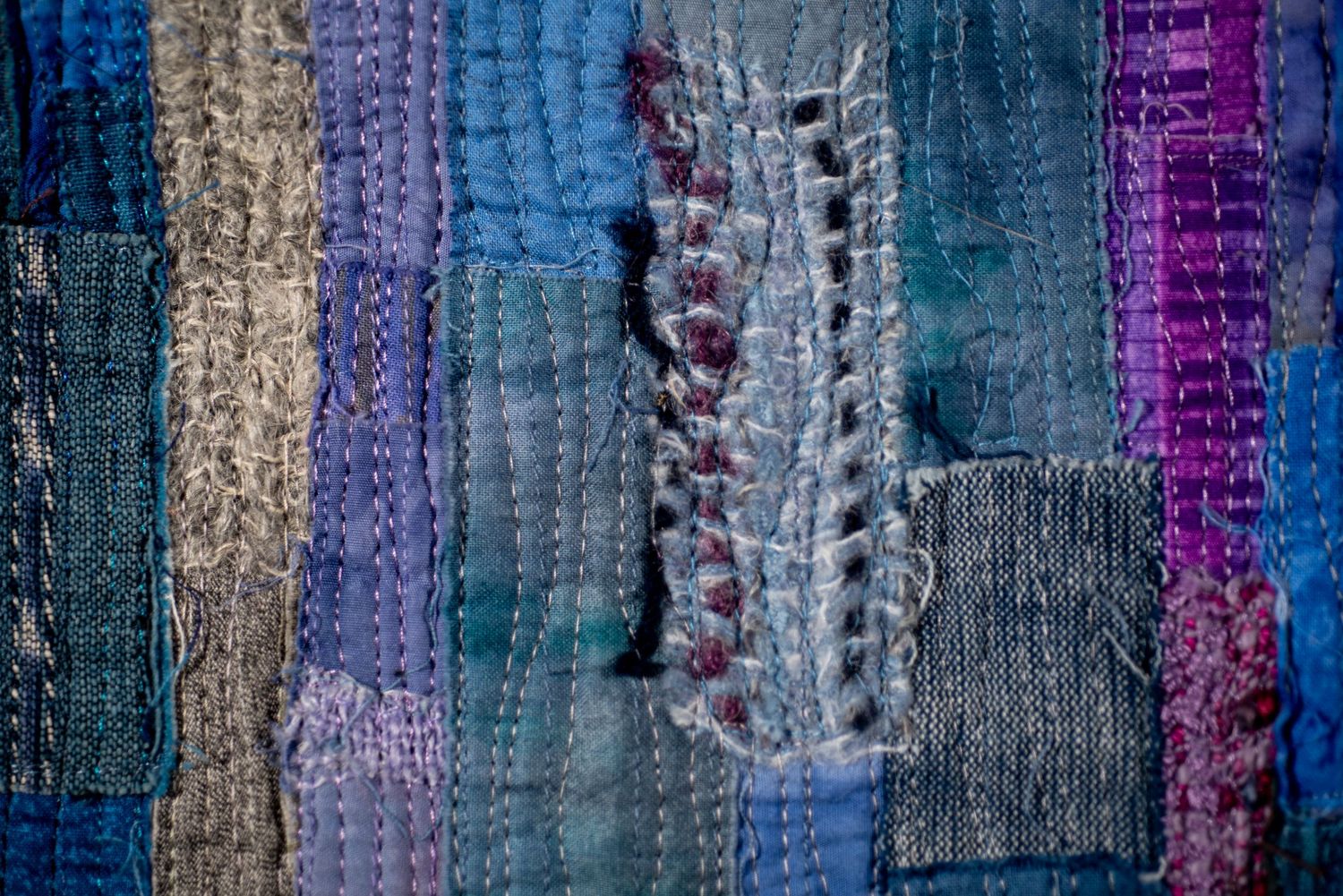 Close-up of patchwork textile with vertical stripes in blue, purple and beige showing detailed stitching and texture.