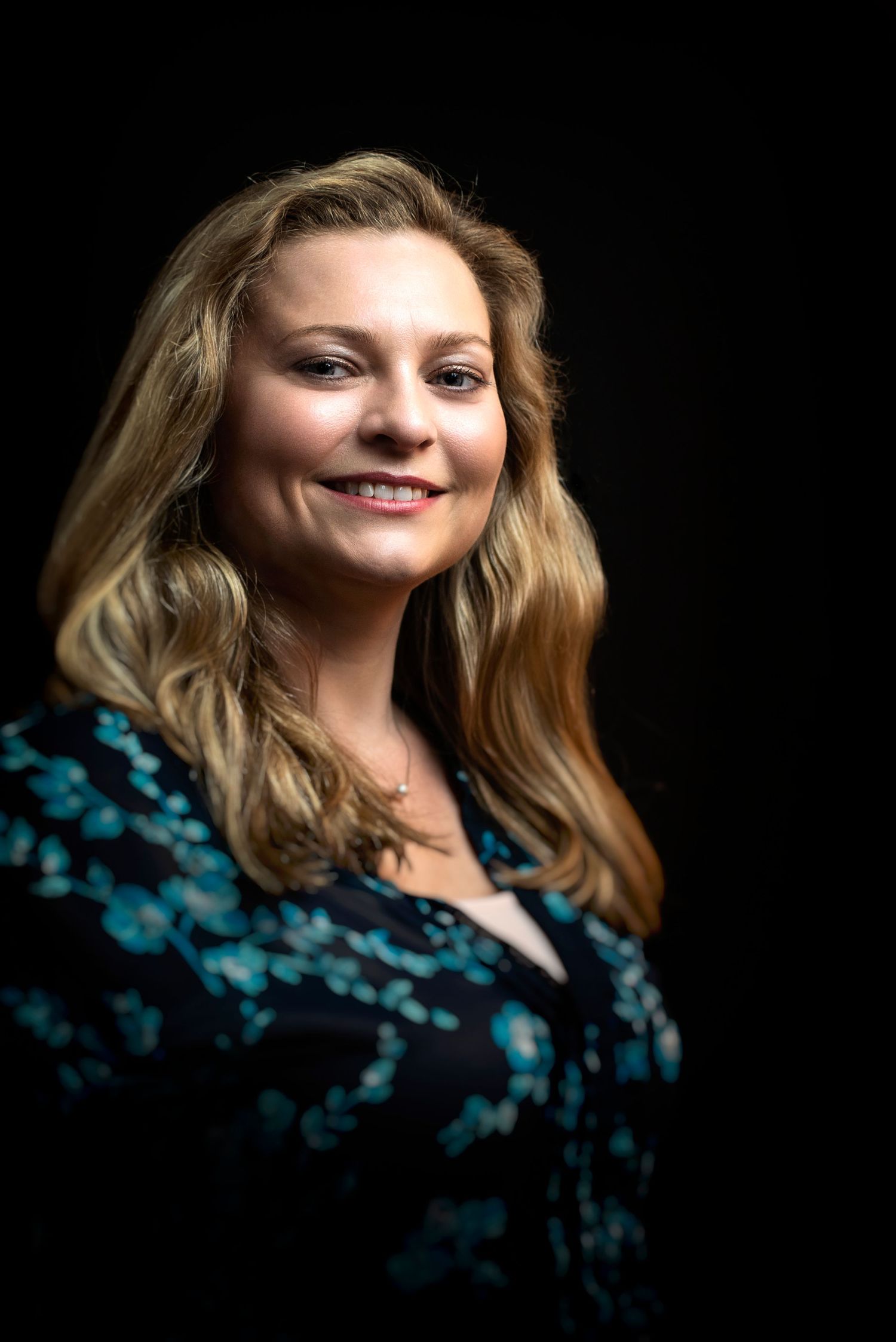 Portrait of a person with blonde wavy hair wearing a dark teal patterned top against a black background.