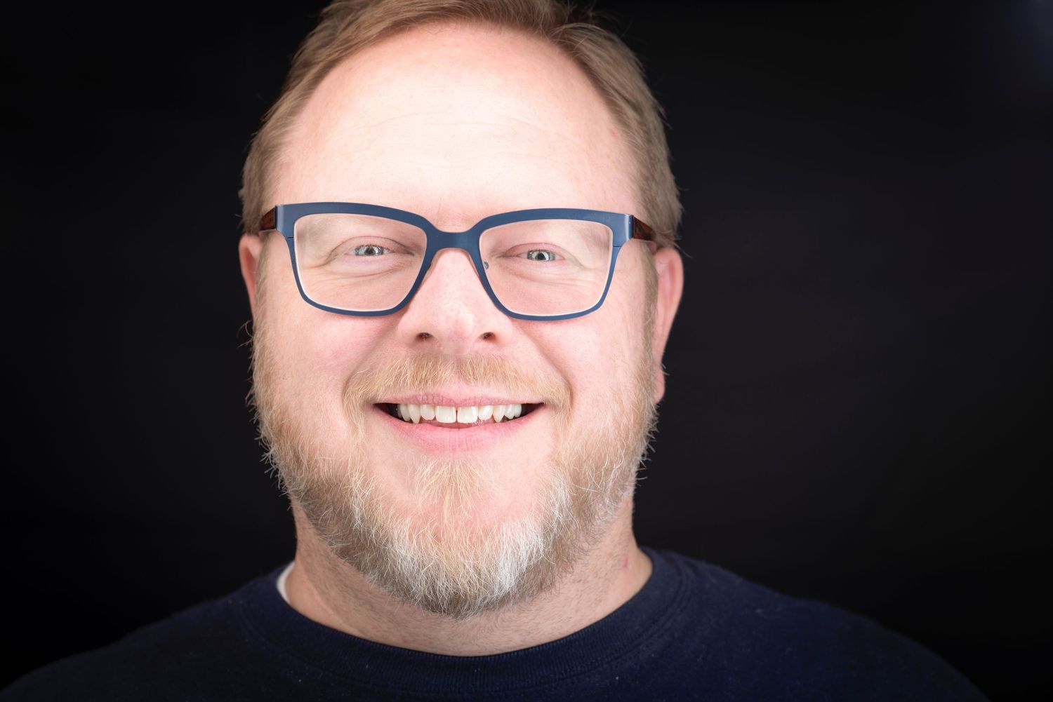Studio portraits of a man wearing glasses and dark clothing against black backdrop.