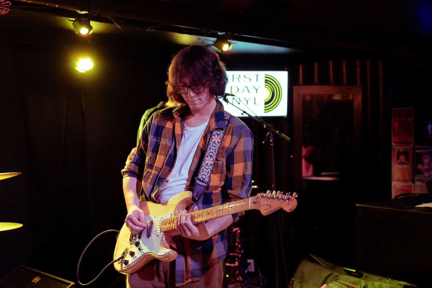 Musicians perform on stage in a dimly lit venue with yellow lighting and musical equipment visible.