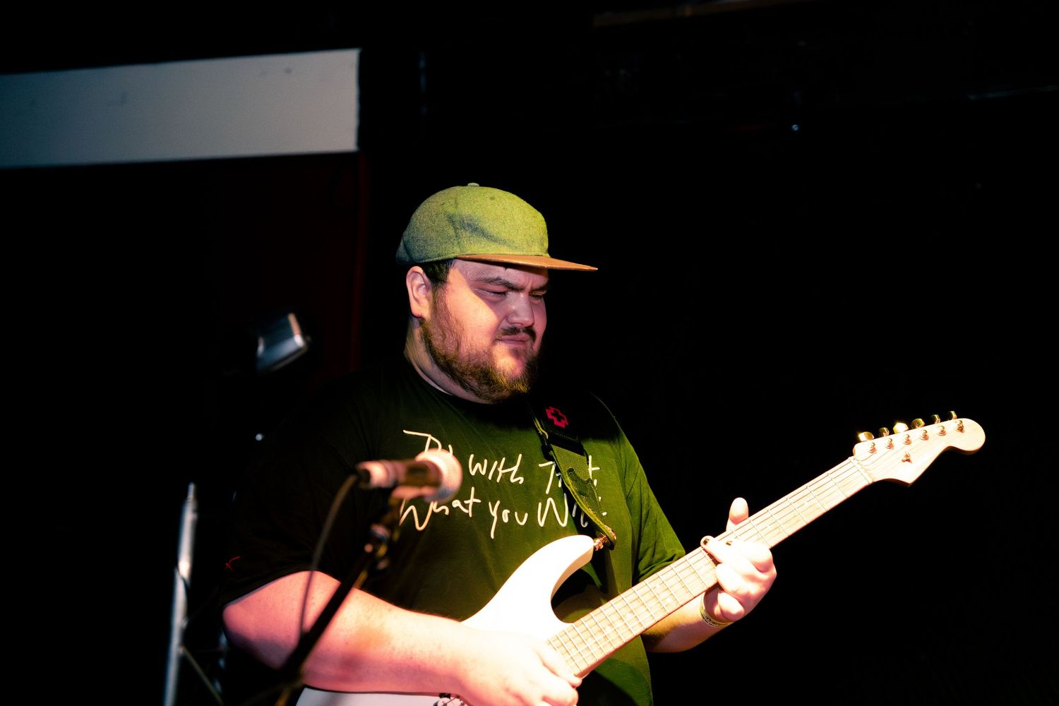 Musician in a green t-shirt and baseball cap plays electric guitar on stage during a live performance.