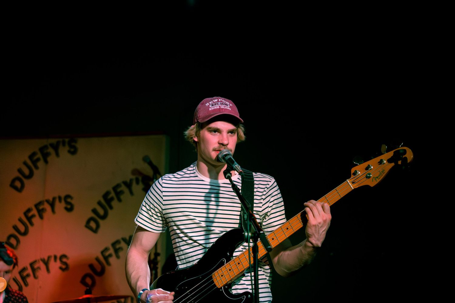 A musician in a striped shirt performs on stage with a bass guitar at Duffy's bar.