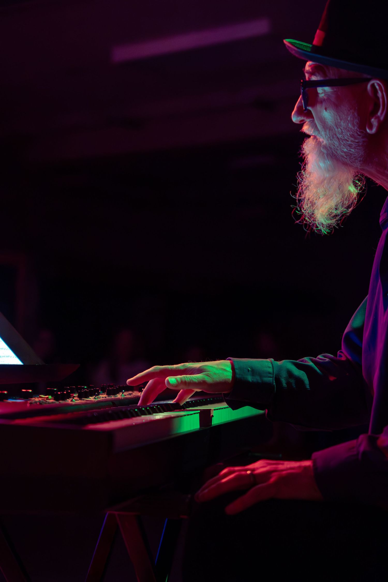 Keyboard player performing on dimly lit stage with purple and green accent lighting.