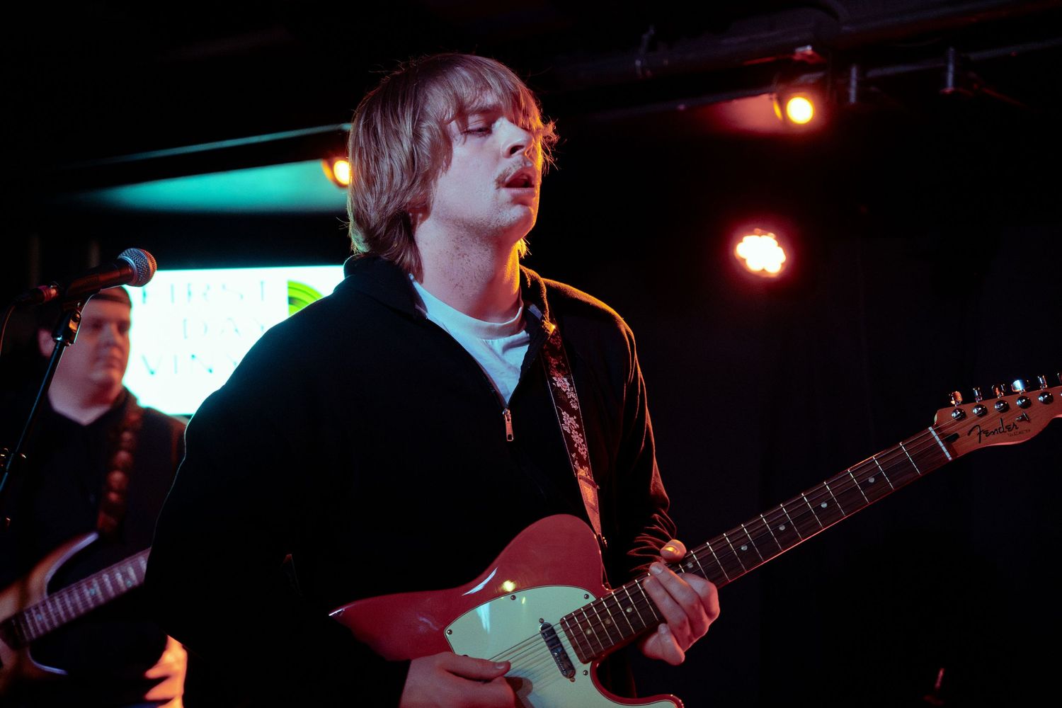 Musician plays electric guitar on stage under red and blue lighting during live performance.