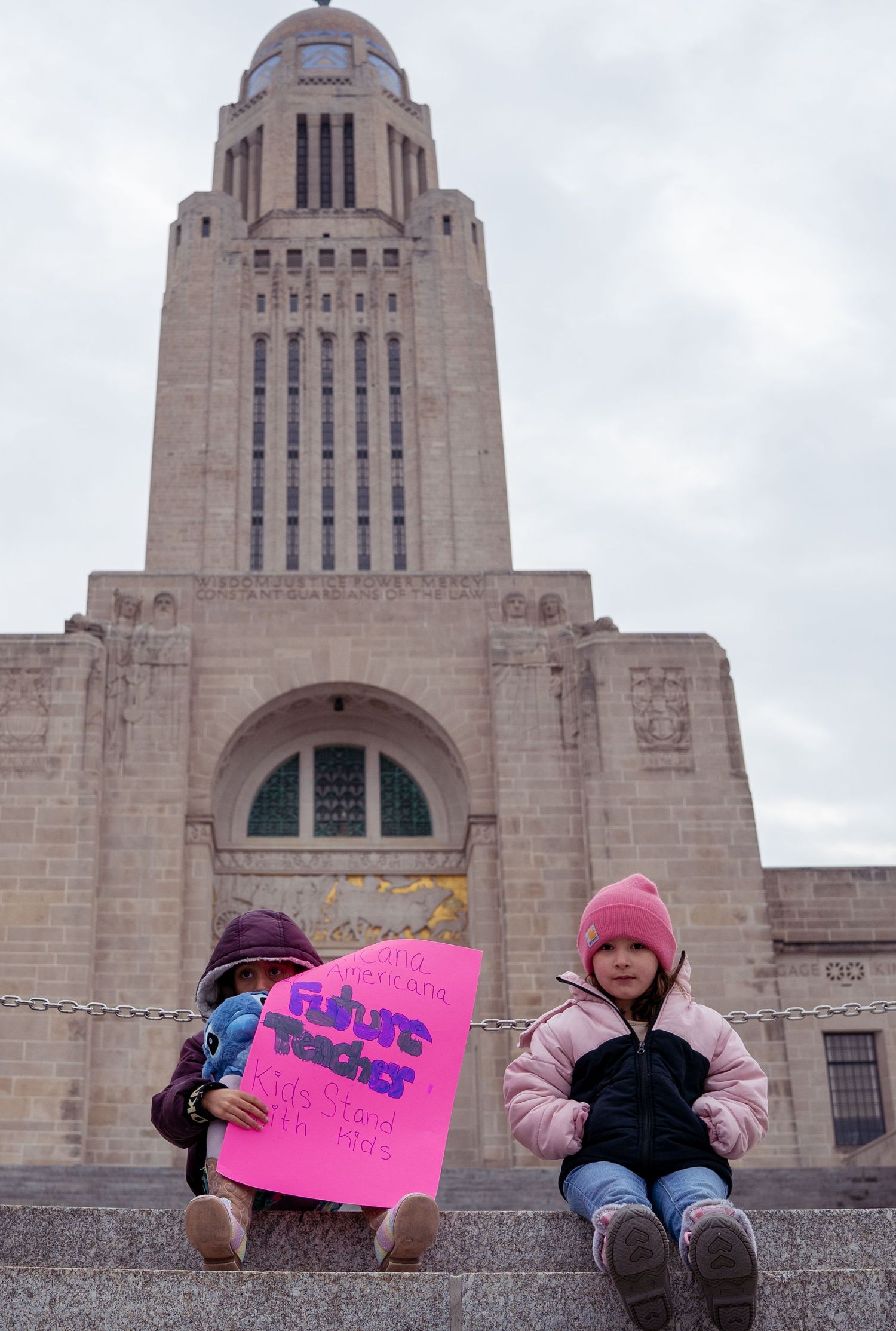 People holding protest signs sit along stone steps of the Nebraska State Capitol building.