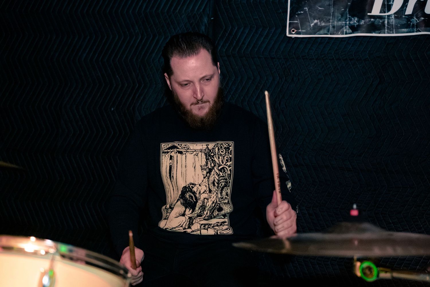 Drummer performs on stage with drum sticks raised while playing a dark-lit drum kit during a live performance.
