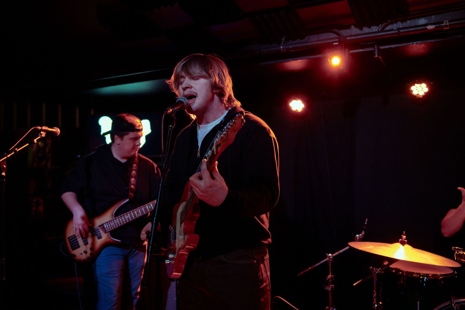 Musicians perform on a dark stage with red and blue lighting while playing guitars at a live concert venue.