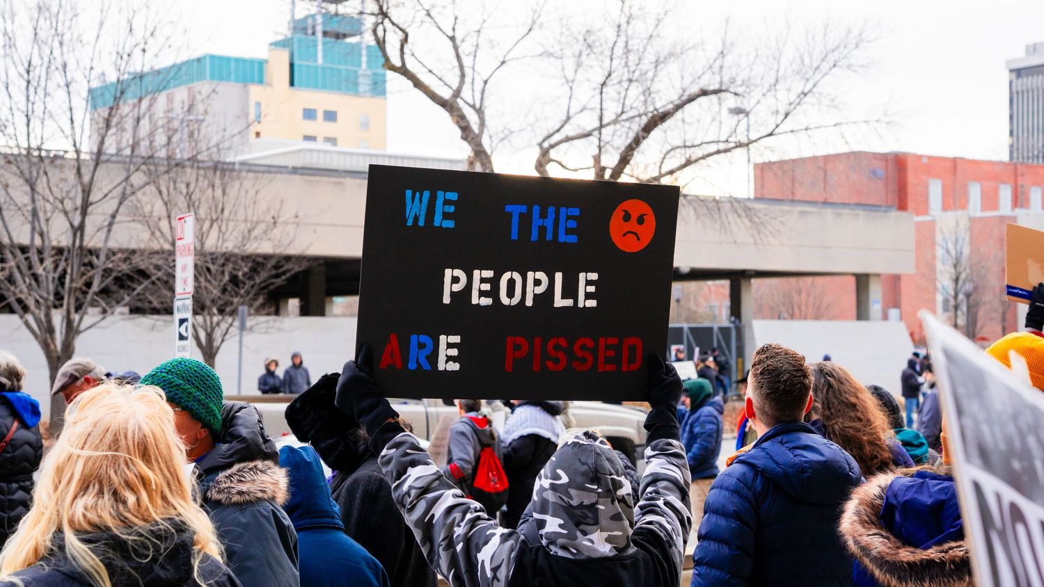Black protest sign reading 'We The People' held up against an urban backdrop.