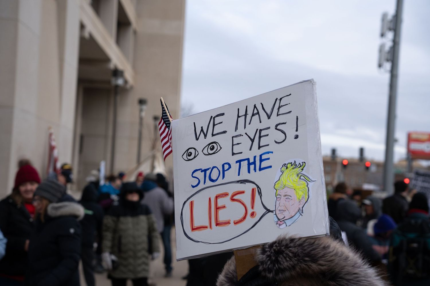 Protest sign reading 'We Have Eyes Stop The Lies' held up in a crowd.