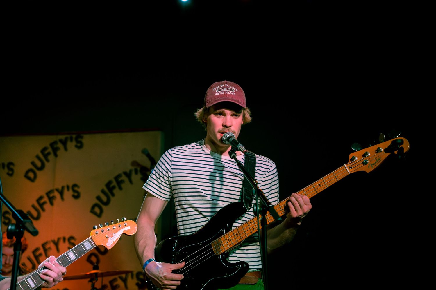 Musician performing on stage at Duffy's bar wearing a striped shirt and playing guitar under green lighting.