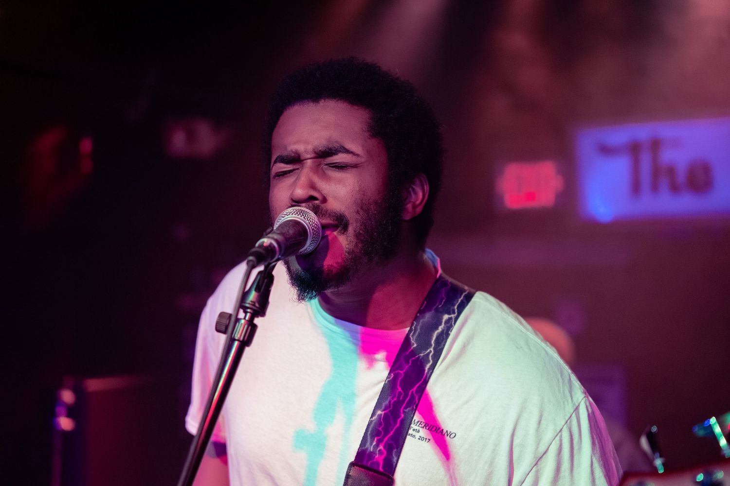 A performer sings passionately into a microphone while playing guitar on stage under red and blue lighting.