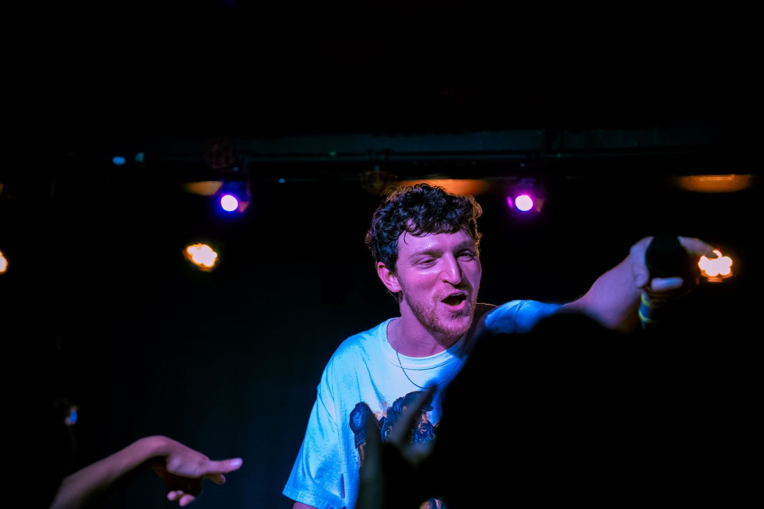 Energetic performer gesturing with arms while singing under colorful stage lights.