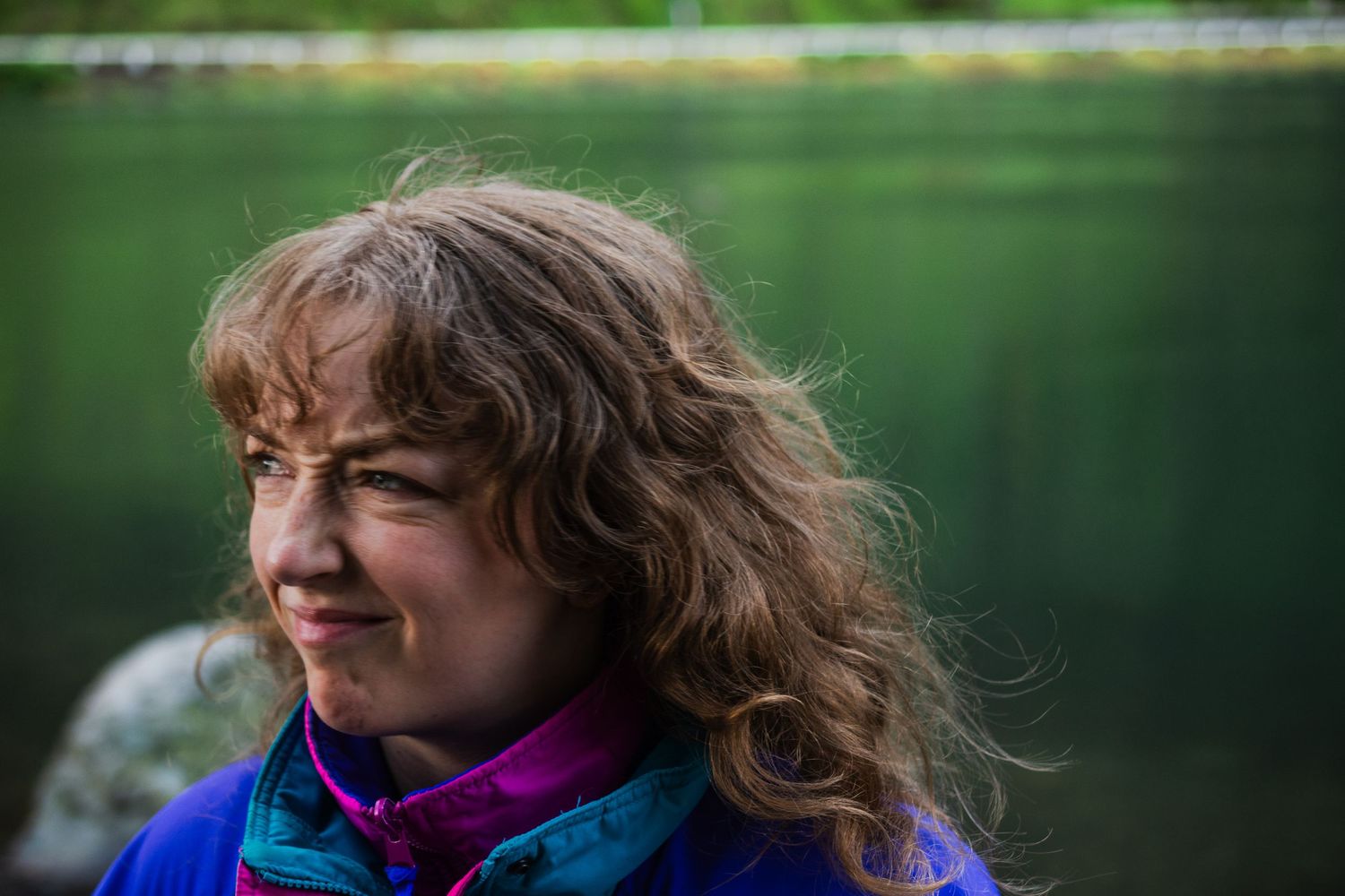 Person with windswept hair enjoying peaceful moment by serene lake with green backdrop.