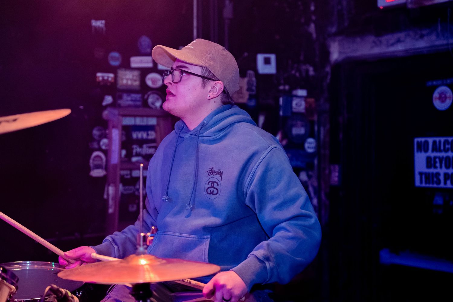 Drummer in a light blue hoodie and baseball cap performs on stage under purple lighting at a music venue.