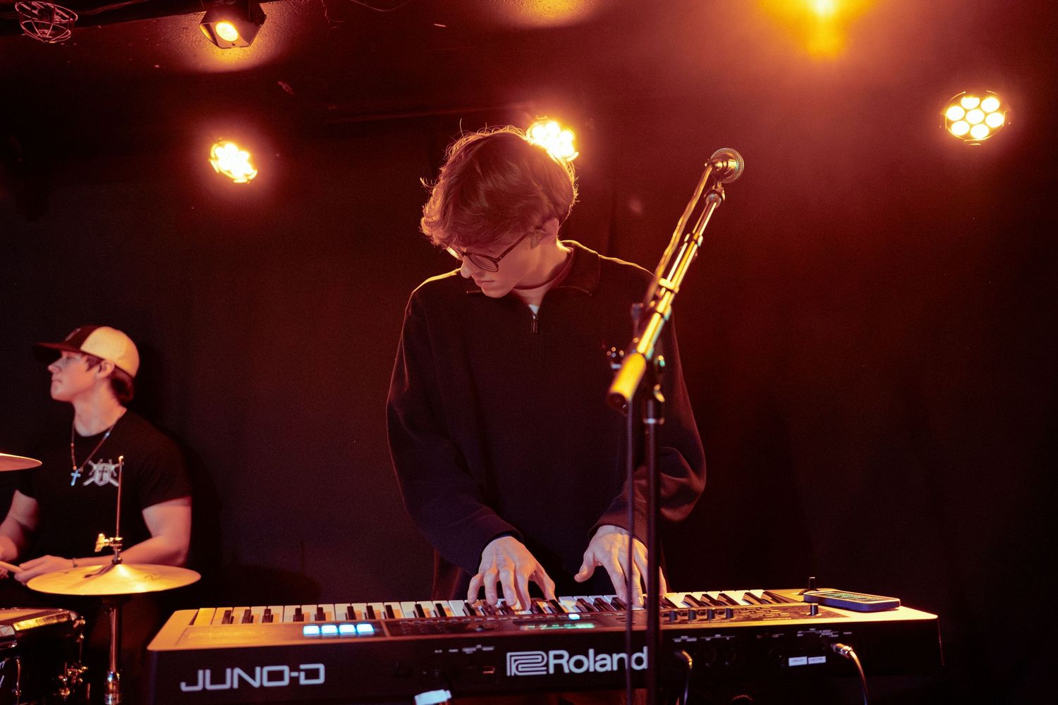 Concert performers play electronic keyboards and instruments on a dark stage illuminated by warm spotlights.