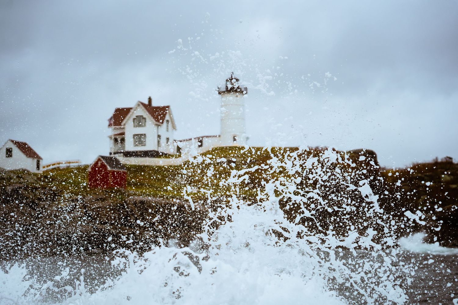 Ocean waves crash against coastal rocks with lighthouse buildings in the background.