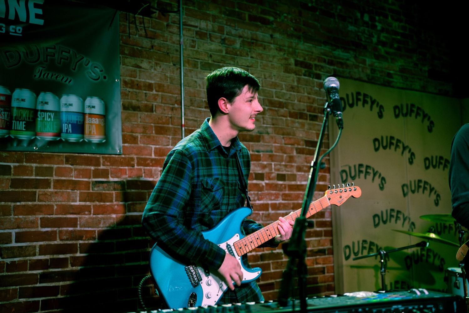 A guitarist performs on stage at a dimly lit venue with brick walls and musical equipment visible in the background.
