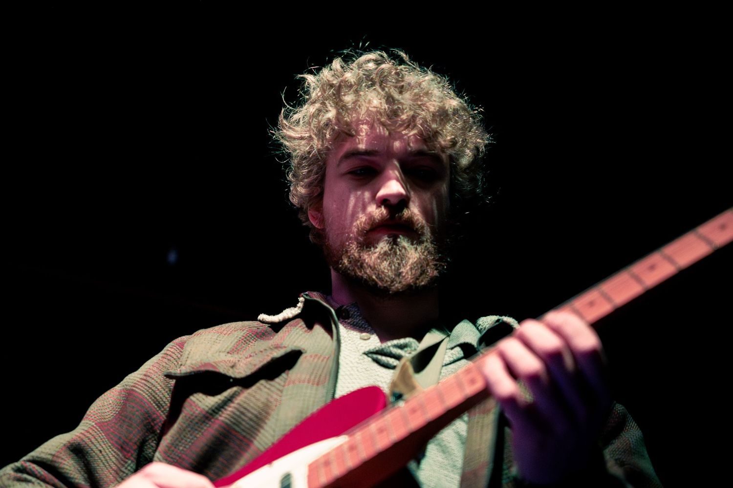 Musician performs passionately on stage with red electric guitar while wearing brown jacket against dark background.