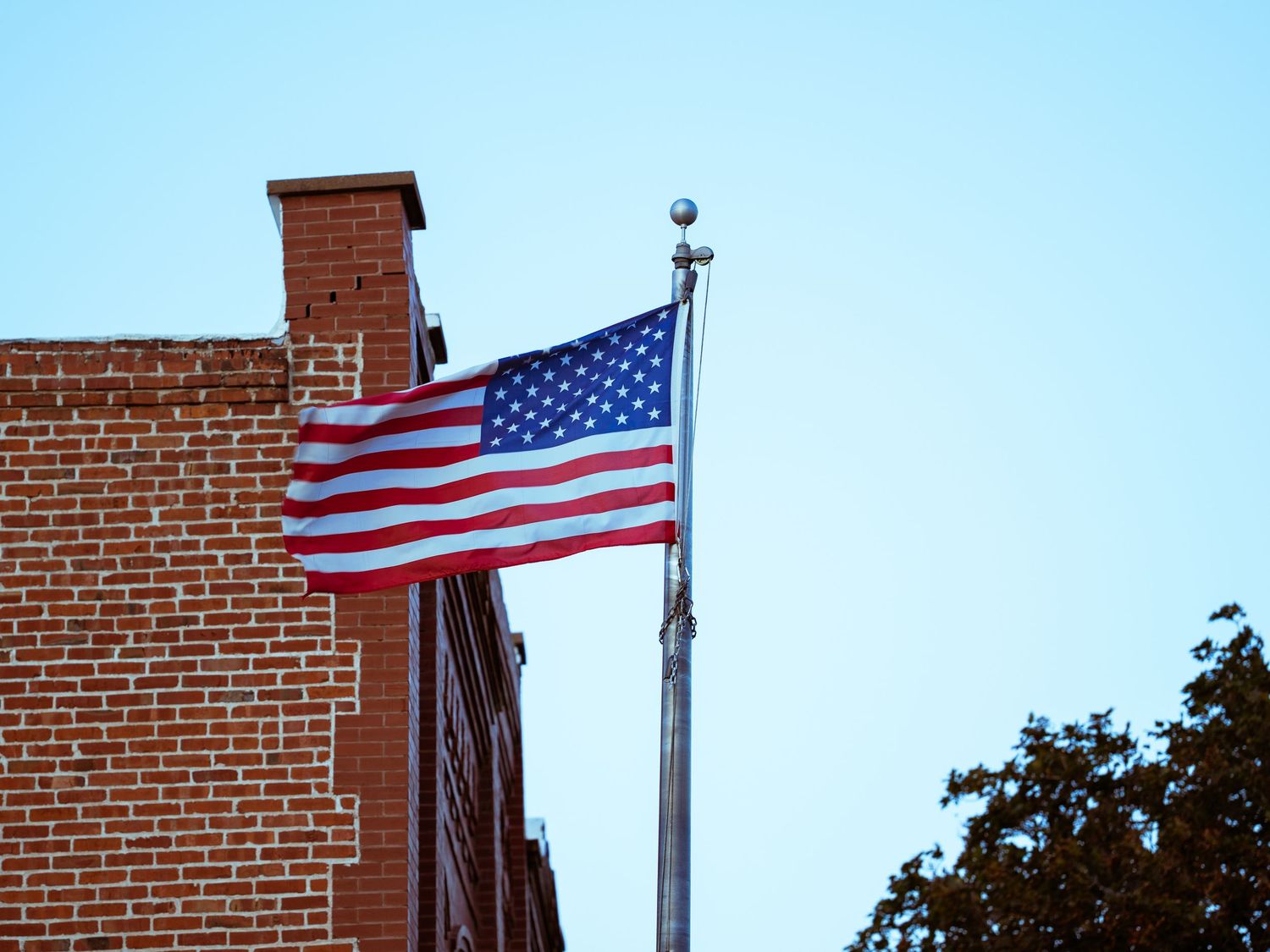 American flag waving against blue sky next to brick building with flagpole.