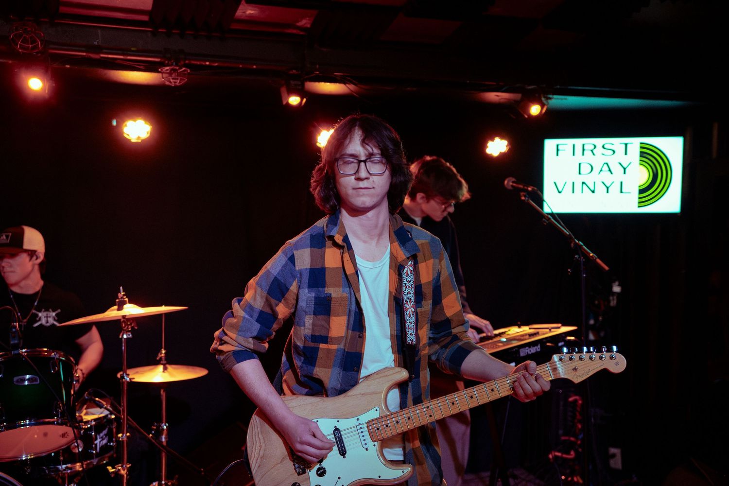 Musicians perform on stage with guitars and drums in a dimly lit venue with red and blue lighting.
