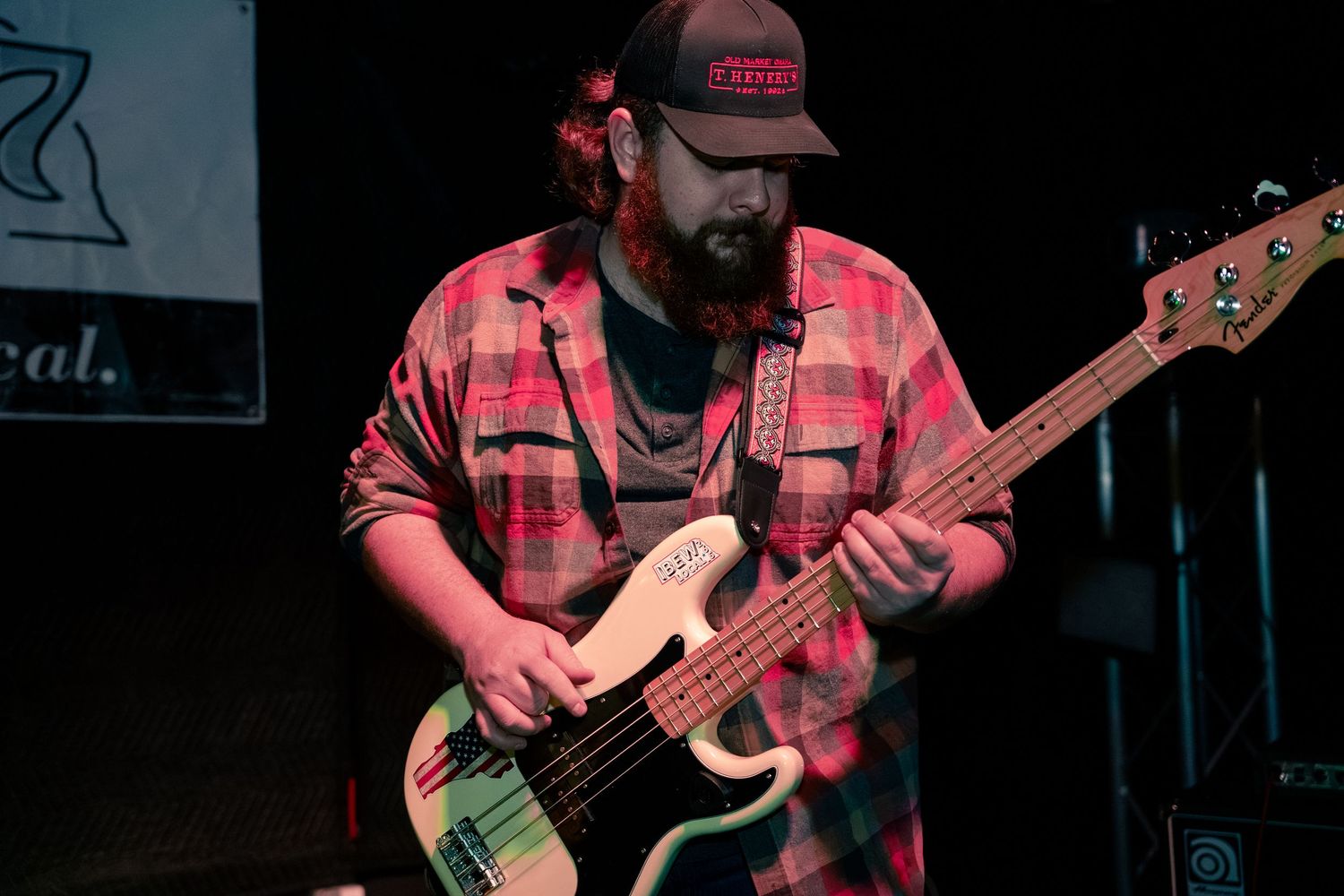 Musician playing bass guitar on stage wearing a red plaid shirt in dark concert lighting.