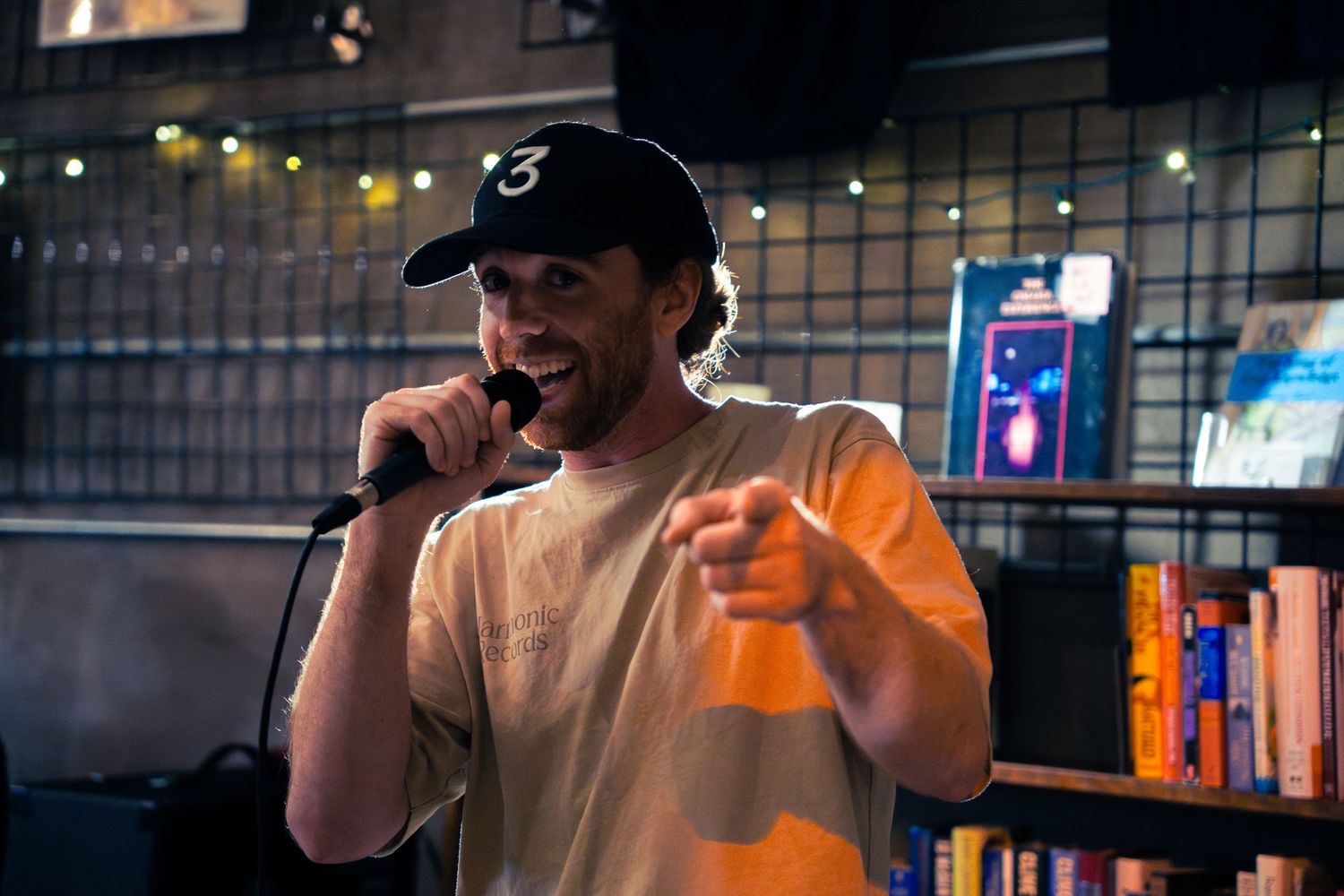 Hip hop artist performing on stage in white t-shirt and baseball cap with neon signs in background.