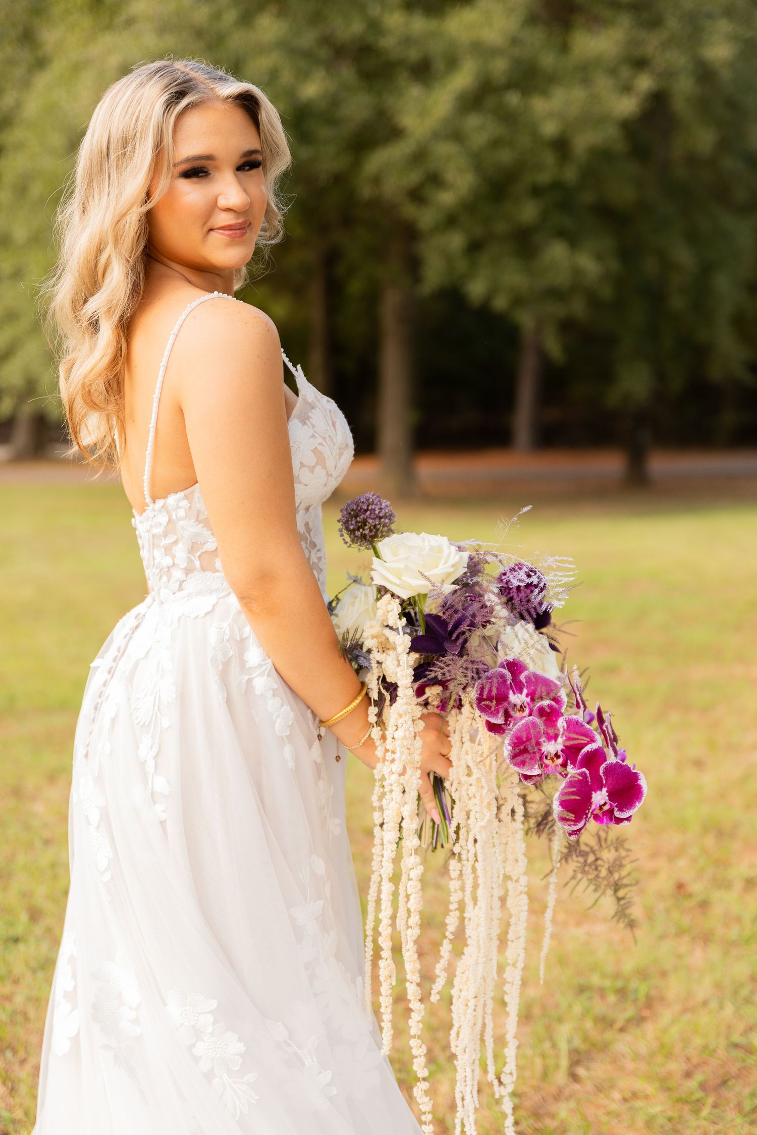 Bride in lace wedding dress holds cascading purple and white bouquet in outdoor setting.