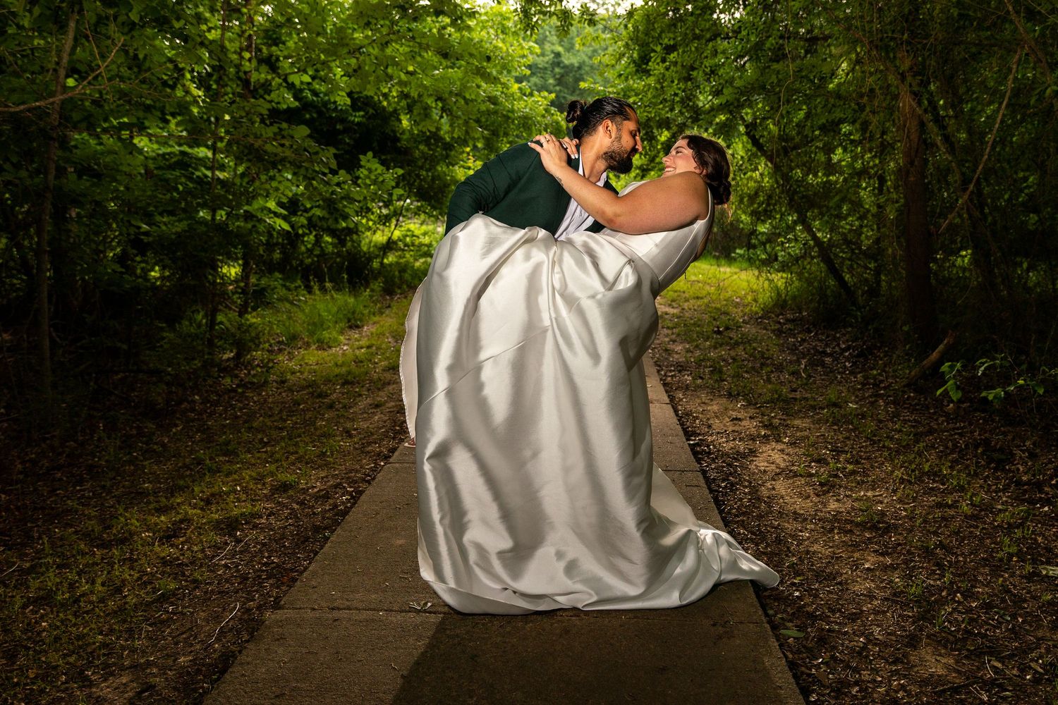 Couple in white attire walks hand in hand down tree-lined path.