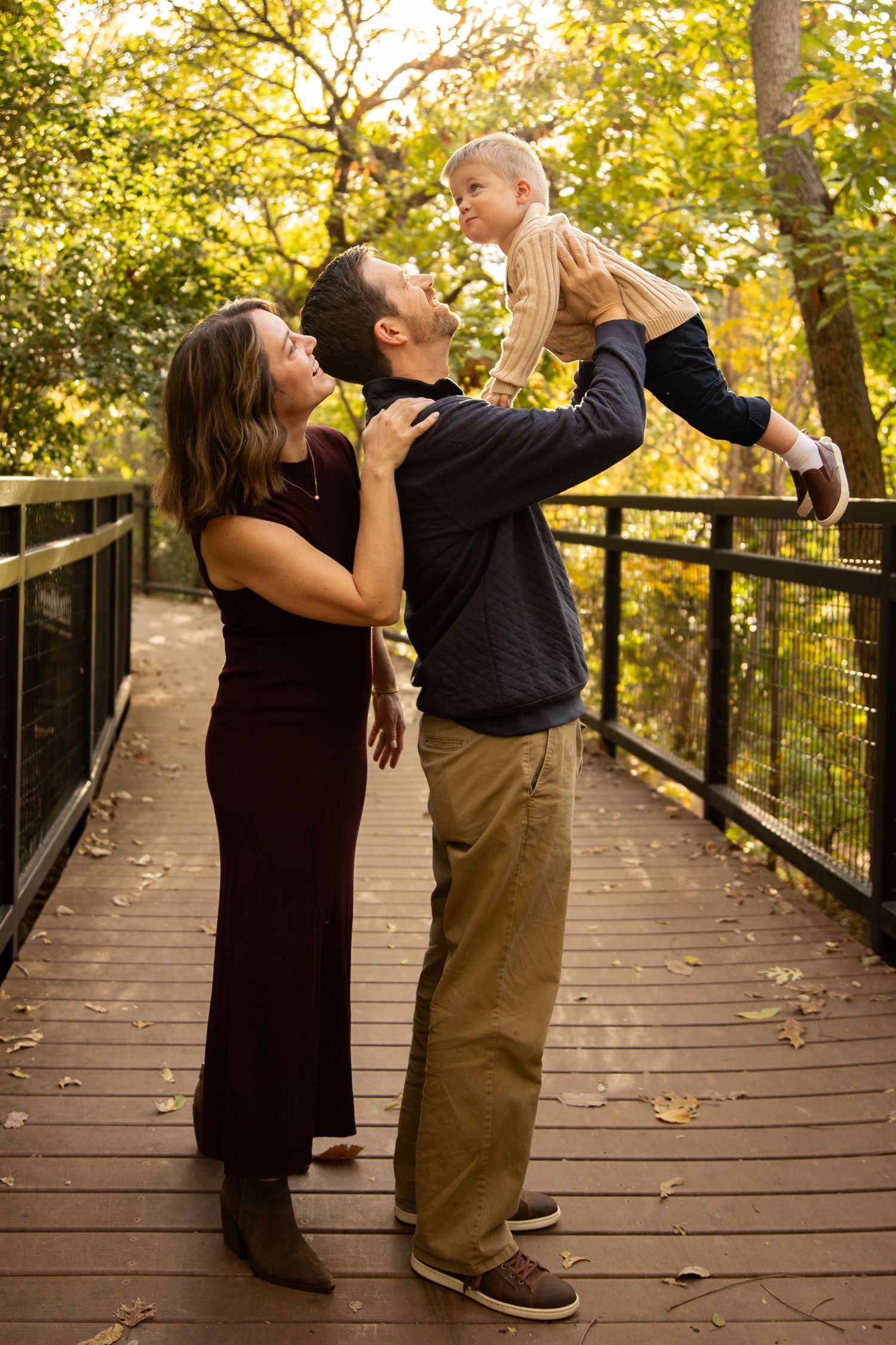 Family members share playful interactions on a wooden bridge surrounded by golden autumn foliage.
